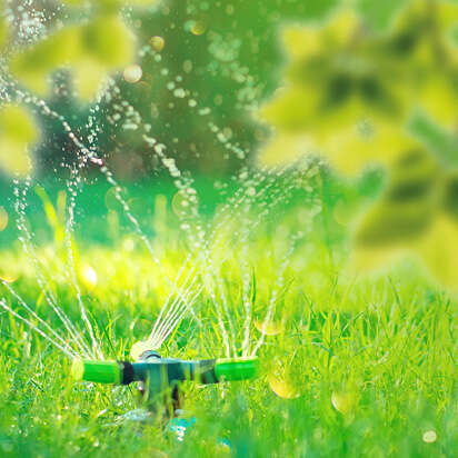 Sprinkler sprüht Wasser auf grünes Gras im Sonnenschein, umgeben von verschwommenen Blättern.